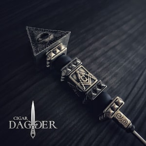 May include: A silver and gold cigar poker with a pyramid-shaped top featuring an all-seeing eye design. The poker has a black rubber grip and several decorative beads with Masonic symbols.