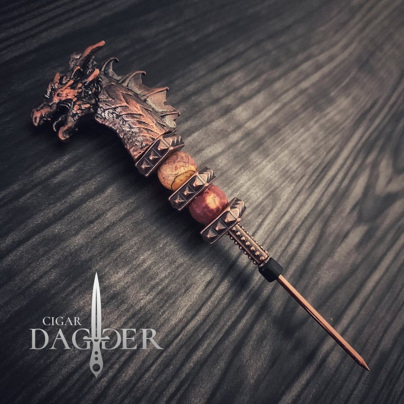 May include: A copper cigar dagger with a dragon head. The handle has gemstone beads and a pointed tip. The text "Cigar Dagger" is below the dagger. The dagger is a decorative accessory.