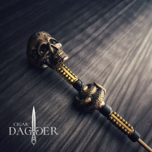 May include: A gold-toned cigar poker with a skull and snake design. The poker is made of metal and has a black handle. The skull is detailed and has a menacing expression. The snake is coiled around the handle and has a realistic appearance. The poker is a unique and stylish accessory for cigar smokers.
