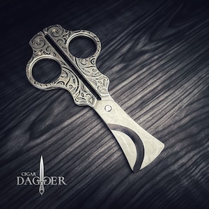 May include: Silver cigar cutter with ornate, swirling patterns etched into the handles. The blade has a curved design with a dark accent. The text "CIGAR DAGGER" is displayed in the lower left corner.
