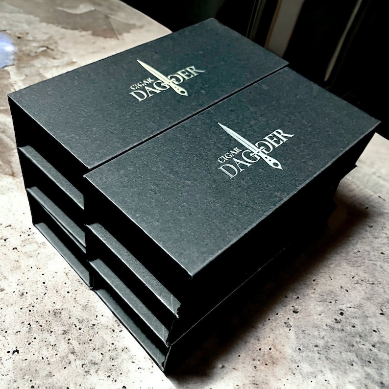 May include: Black boxes with the text "CIGAR DAGGER" and a dagger logo in white. The boxes are stacked, revealing multiple compartments. The boxes appear to be made of a textured material.