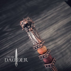 May include: A decorative cigar dagger with a bronze-coloured metal handle and a detailed lion's head design. The handle features a textured pattern and is adorned with orange and red beads. The text "Cigar Dagger" is displayed in the lower left corner.
