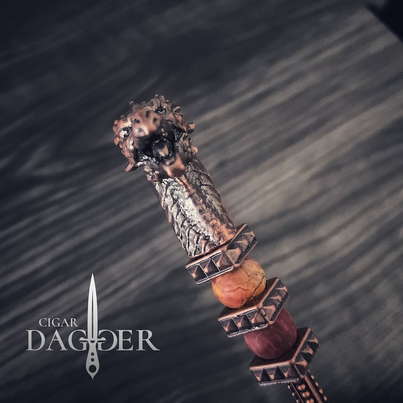 May include: A decorative cigar dagger with a bronze-coloured metal handle and a detailed lion's head design. The handle features a textured pattern and is adorned with orange and red beads. The text "Cigar Dagger" is displayed in the lower left corner.