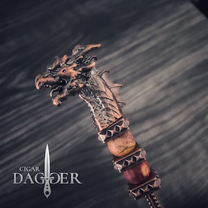 May include: A bronze cigar dagger with a dragon head. The handle has metal bands and natural stone beads. The text "Cigar Dagger" is printed below the dagger. The overall design has a medieval theme.