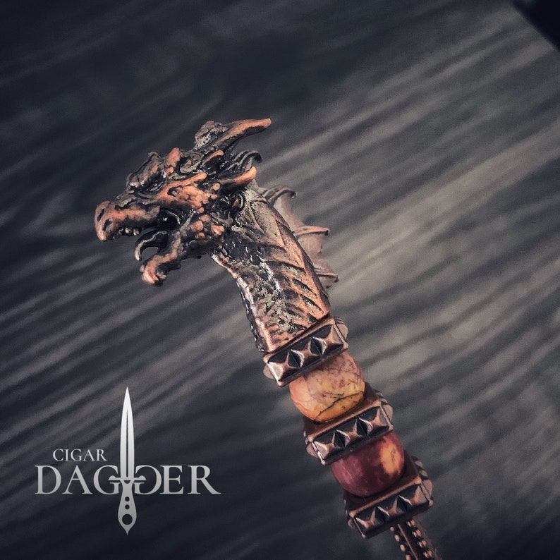 May include: A bronze cigar dagger with a dragon head. The handle has metal bands and natural stone beads. The text "Cigar Dagger" is printed below the dagger. The overall design has a medieval theme.