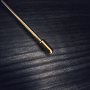 May include: A gold-coloured lapel pin with a rounded tip, set against a dark, textured background. The pin is straight with a cylindrical design, reflecting light. The overall aesthetic is minimalist and elegant, suitable for formal wear.