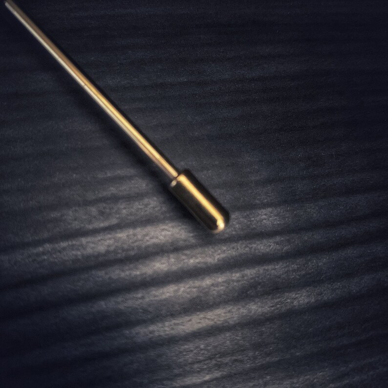 May include: A gold-coloured lapel pin with a rounded tip, set against a dark, textured background. The pin is straight with a cylindrical design, reflecting light. The overall aesthetic is minimalist and elegant, suitable for formal wear.
