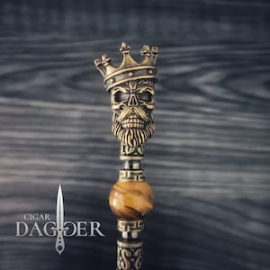 Pode incluir: Um crânio dourado com barba usando uma coroa, com uma pedra de olho de tigre e uma faixa decorativa abaixo. O crânio está em um fundo escuro.