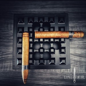 May include: Two brown cigars resting on a black gridded tray. The tray has a raised edge and is made of a textured material. The cigars are both lit and have a white band around them.