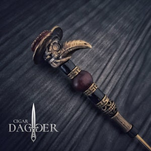 May include: A gold and black cigar poker with a plague doctor skull design. The poker has a red gemstone on the top and a wooden bead in the middle.