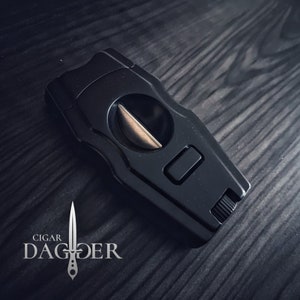 May include: Black cigar cutter with a silver blade and a push button. The cutter is on a dark wood surface.