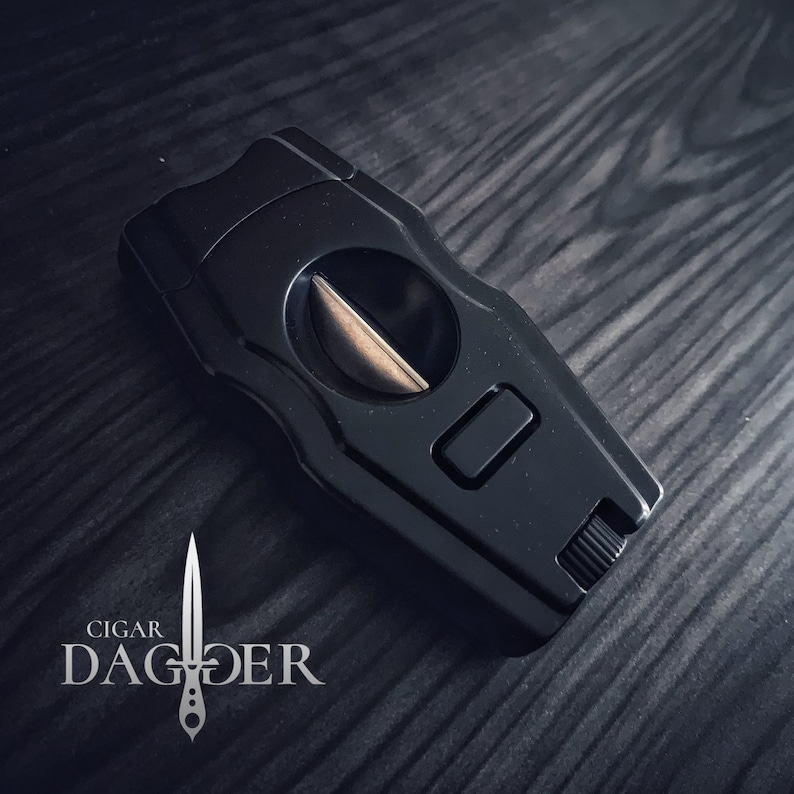 May include: Black cigar cutter with a silver blade and a push button. The cutter is on a dark wood surface.