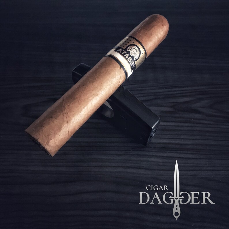 May include: A brown cigar with a gold band that says "ATABEY" on a black cigar holder. The cigar is on a dark wood surface.