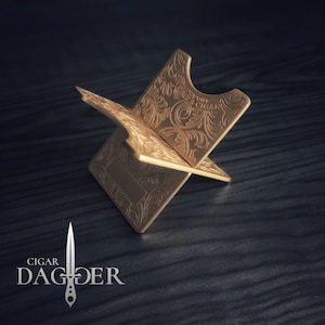 May include: A gold-colored metal cigar stand with an ornate design. The stand is shaped like a cross and has a small, round indentation in the center for holding a cigar.