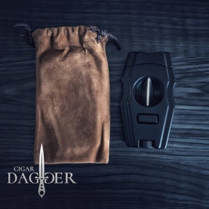 May include: A black cigar cutter with a silver blade and a brown velvet pouch. The cutter has a  "CIGAR DAGGER" logo on the pouch.