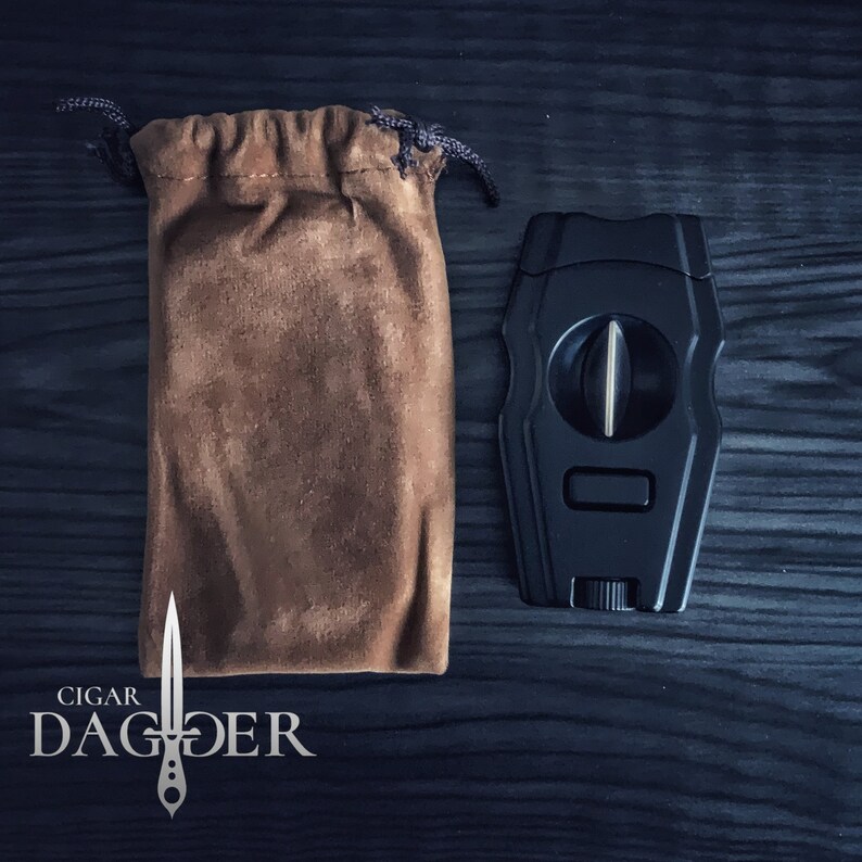 May include: A black cigar cutter with a silver blade and a brown velvet pouch. The cutter has a  "CIGAR DAGGER" logo on the pouch.