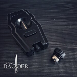 May include: A black cigar cutter with a guillotine blade and a push button release. The cutter is made of metal and has a sleek, modern design. The cutter is sitting on a dark wooden surface. The brand name "CIGAR DAGGER" is visible in the lower left corner of the image.