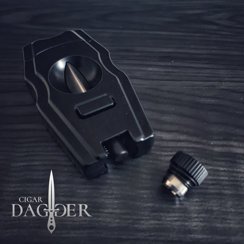 May include: A black cigar cutter with a guillotine blade and a push button release. The cutter is made of metal and has a sleek, modern design. The cutter is sitting on a dark wooden surface. The brand name "CIGAR DAGGER" is visible in the lower left corner of the image.