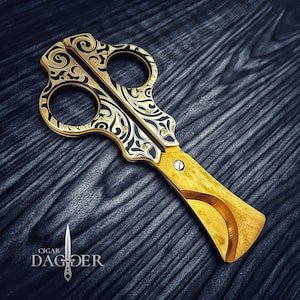May include: Gold cigar cutter with a black and gold floral design. The cutter has a curved blade and a small hole for cutting the end of a cigar.