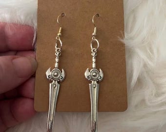 May include: A pair of silver sword-shaped dangle earrings with a detailed design. The earrings are hanging on a brown card.