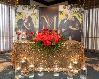 May include: A gold sequined tablecloth with a large red floral arrangement in the center. There are clear glass cylinders with candles on the table. The backdrop is a black and gold banner with the letter "M" in gold and two photos of a couple.