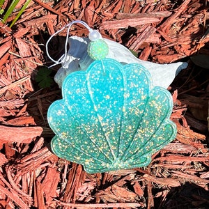 May include: A teal blue seashell ornament with gold glitter. The seashell has a white string attached to it with a white bead and a green glitter bead.