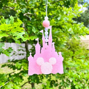 May include: Pink glitter castle car air freshener with a white Mickey Mouse head in the center. The castle has white glitter accents and is hanging from a white string with pink and white beads.