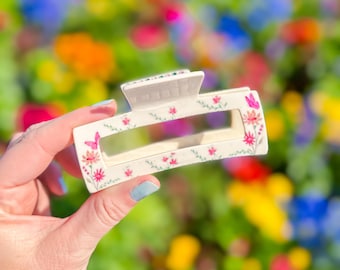 May include: White plastic hair claw with a floral pattern featuring pink, red, and green flowers. The claw is in focus, while the background is blurred and shows a field of colorful flowers.