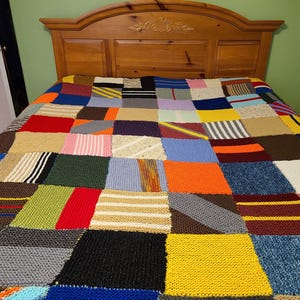 Könnte beinhalten: Handgefertigte Patchwork-Decke mit gestrickten Quadraten und Rechtecken in verschiedenen Farben, darunter Rot, Blau, Gelb und Grün. Die Decke liegt auf einem hölzernen Bettrahmen.