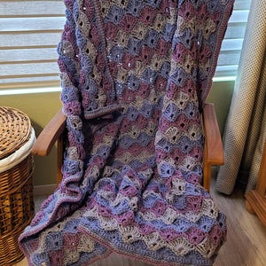 May include: A handmade crochet blanket draped over a wooden chair. The blanket features a repeating pattern in shades of purple, blue, and gray. The blanket's edges are finished with a contrasting border. The chair is made of wood and has a natural finish.
