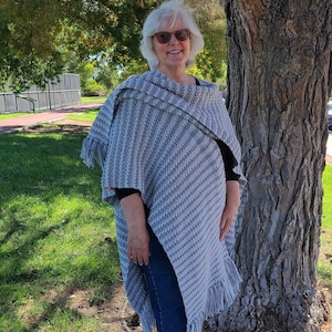 Könnte beinhalten: Eine Frau trägt einen grauen und weißen gestreiften gehäkelten Poncho mit Fransen. Sie steht vor einem Baum und trägt eine Sonnenbrille.