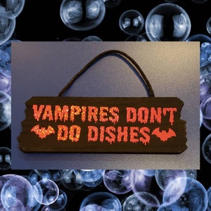 May include: A rectangular wooden sign with the text "VAMPIRES DON'T DO DISHES" in orange glitter lettering. A bat graphic is featured below the text. The sign has a black background and a black string for hanging.