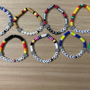 May include: Eight colorful beaded bracelets with white beads spelling out names and numbers. The bracelets feature a checkered pattern on the center bead.