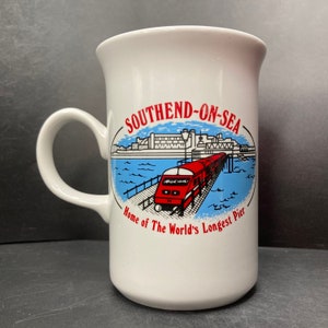Może przedstawiać: Biały kubek ceramiczny z czerwoną, białą i niebieską ilustracją pociągu na molo. Tekst "Southend-on-Sea" jest czerwony, a tekst "Home of The World's Longest Pier" jest czarny.