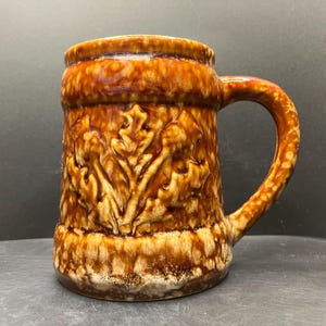 May include: A brown ceramic beer mug with a handle. The mug has a textured surface with a raised leaf design. The stein has a glossy finish and a wide base. The handle is rounded and attached to the side of the mug.