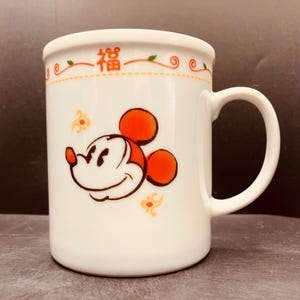 May include: A white ceramic mug featuring an orange graphic of Mickey Mouse's head. The top rim is decorated with an orange border, small green leaves, and the Chinese character for good fortune, 福. Additional orange floral accents surround the character.