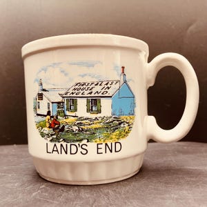May include: A white ceramic mug featuring a colourful illustration of a building labelled FIRST & LAST HOUSE IN ENGLAND and the text LAND'S END. The design shows a small house with green shutters, a chimney, and two figures sitting on the grass.