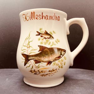 Peut inclure: Une tasse en céramique de couleur crème portant le mot Killeshandra en écriture brune sur le dessus. Le corps présente une illustration détaillée de deux poissons nageant parmi des plantes aquatiques vertes et des rochers bruns à la base.
