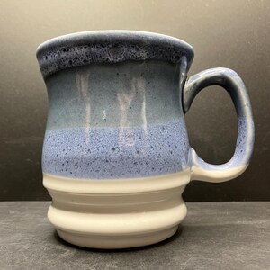 May include: A handmade ceramic mug with a blue and white glaze. The mug features a dark blue rim, transitioning to lighter shades of blue, and a white base. The handle is also blue and white.