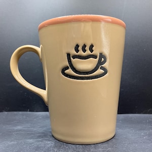 May include: Tan ceramic coffee mug with a brown handle and rim. A black graphic of a steaming coffee cup is on the front. The mug is designed for hot beverages.