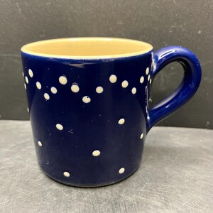 May include: A ceramic mug with a navy blue exterior and a tan interior. The mug is decorated with white polka dots. It has a curved handle and is suitable for hot or cold beverages.