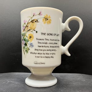 May include: White ceramic mug with a pedestal base and heart-shaped handle. The mug features a painted bird, flowers, and the text "THE SONG OF LIFE" with a poem. The mug is a decorative item.