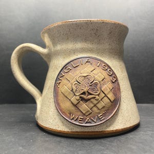 Pode incluir: Uma caneca de cerâmica bege com uma alça. A caneca apresenta um emblema circular castanho com o texto "ANGLIA 1999" e "WEAVE" em torno de um desenho de trevo. A caneca tem uma textura salpicada.