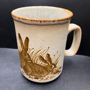 May include: A ceramic mug with a beige speckled finish and a brown rim. The mug features a brown illustration of two rabbits in a grassy field. The handle is a matching beige color.