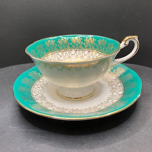 Peut inclure: Un service à thé vintage composé d'une tasse et d'une soucoupe. La tasse et la soucoupe sont blanches avec une bordure turquoise et des détails dorés. La tasse a une anse incurvée et repose sur un petit piédestal.