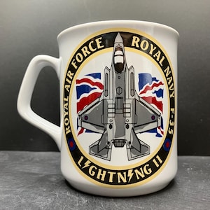 May include: White ceramic mug with a design featuring a grey F-35 Lightning II fighter jet, and the text "Royal Air Force Royal Navy F-35 Lightning II". The mug has a large handle and is suitable for hot beverages.