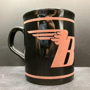 May include: Black coffee mug with a handle, featuring a salmon-colored winged design and the letter "B". The mug has a salmon-colored stripe near the top and bottom. The mug is likely ceramic.