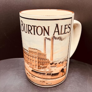 May include: A cream-coloured ceramic mug featuring a brown printed illustration of a large industrial brewery complex with two tall smokestacks emitting smoke. The text BURTON ALES is printed in a bold, dark brown serif font above the factory image.