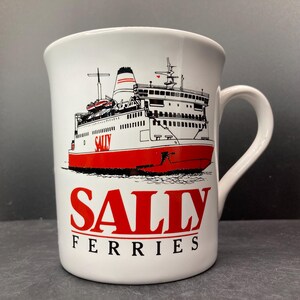 May include: White ceramic mug featuring a red and black illustration of a ferry boat. The word "SALLY" is printed in red, with "FERRIES" below it in black. The mug has a curved handle.