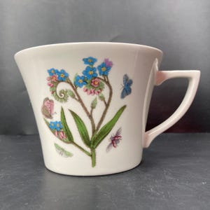 Peut inclure: Une tasse à thé en céramique blanche avec un motif floral composé de fleurs bleues myosotis, de feuilles vertes et de papillons roses et bruns. La tasse a un bord évasé et une anse.